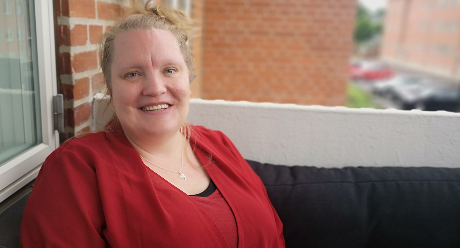 Portrætbillede af social- og sundhedshjælper Tanja Rasmussen, der sidder på en sofa på en altan i en rød murstensbygning og smiler til kameraet.