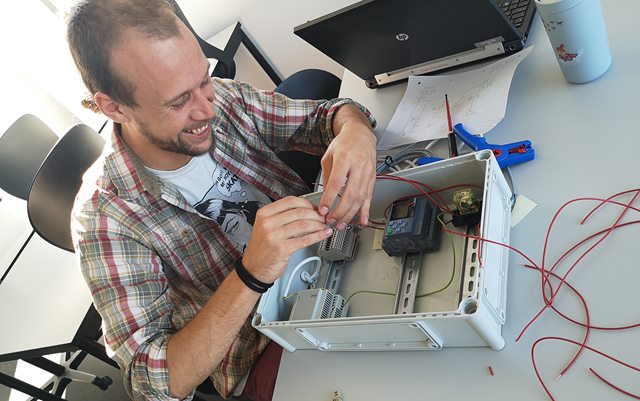 Automationsteknolog-studerende Anders Povlsen sidder ved et skrivebord og arbejder på en eltavle med en lille skruetrækker. Han er iført en ternet skjorte og smiler, mens han ser ned på komponenterne. 