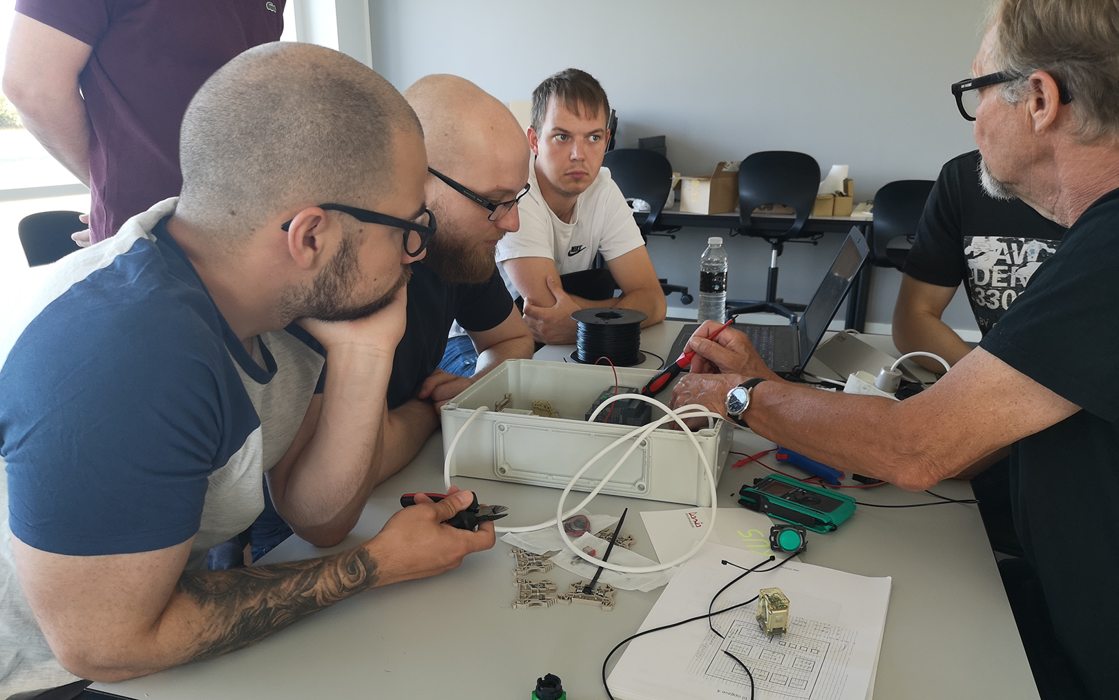 Underviser Lars Dam sidder ved en eltavle på et skrivebord med en skruetrækker i hånden. Fem studerende er samlet tæt omkring eltavlen, imens underviseren fortæller. 