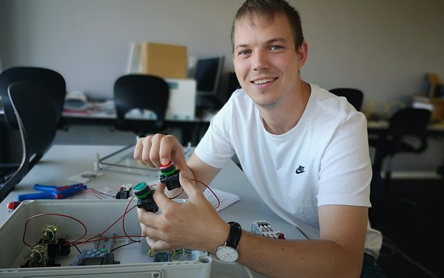 Automationsteknolog-studerende Mathias Mortensen sidder ved et skrivebord med nogle elkomponenter i hånden og smiler til kameraet. Han er iført hvid t-shirt.
