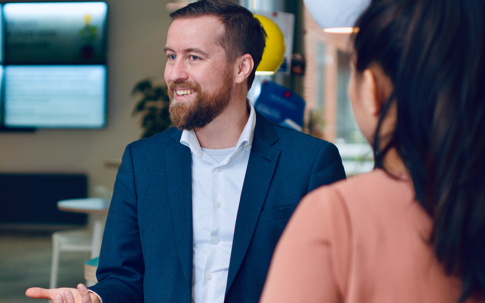 Underviser Claus Grand Bang i samtale. Han er iført hvid skjorte og blå jakke og gestikulerer med hånden, mens han smiler. På billedet ses også en mørkhåret kvinde bagfra.