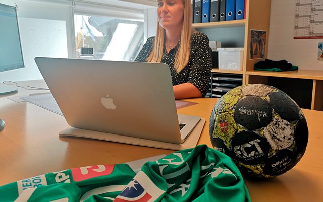 Rikke Sørensen sidder bag computer ved skrivebord. På skrivebordet ligger håndbold og hold-t-shirt. 
