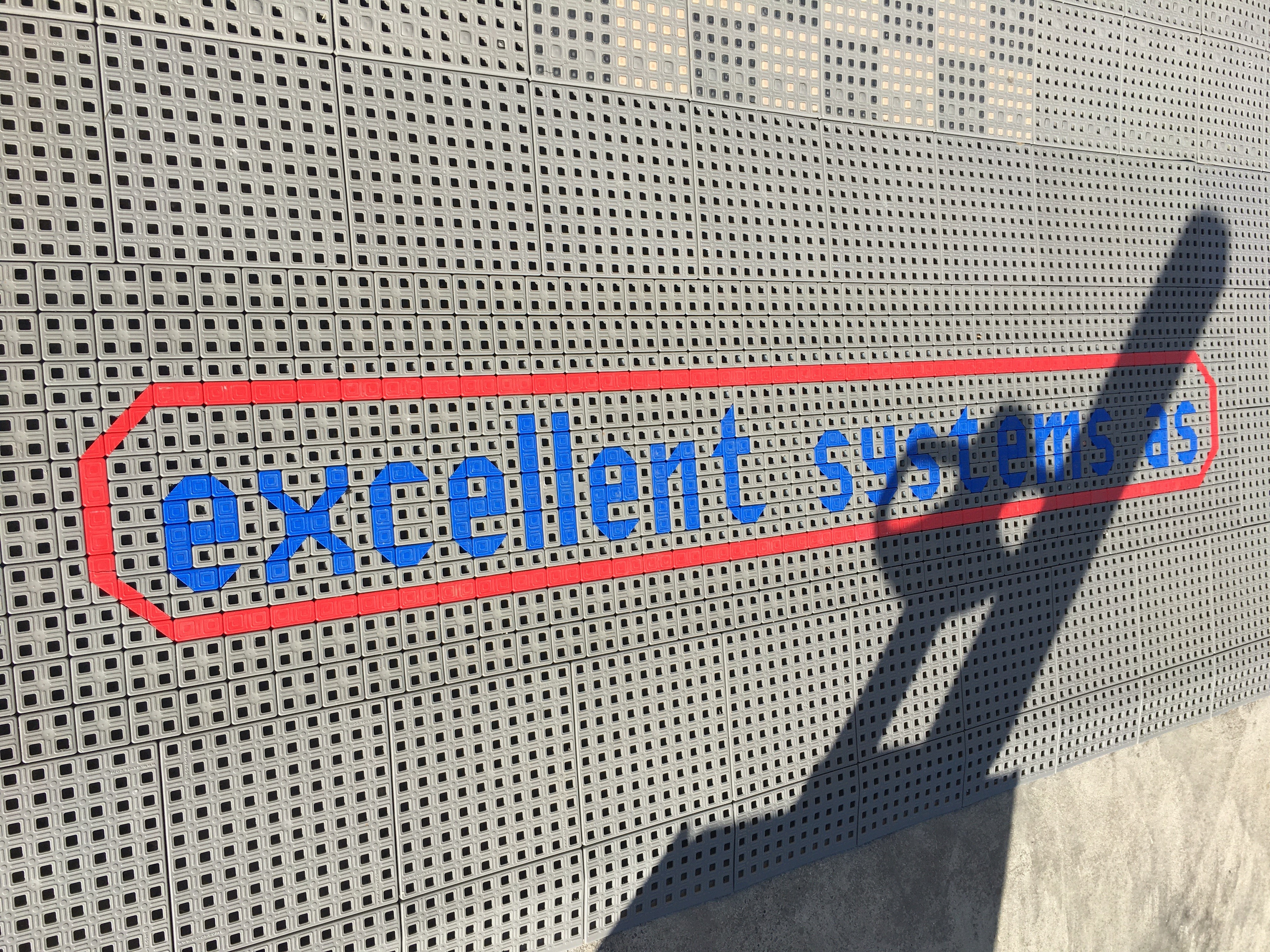 Et aflangt stykke gråt skridsikkert gulv med firmanavnet 'Excellent Systems' skrevet med lyseblå bogstaver.