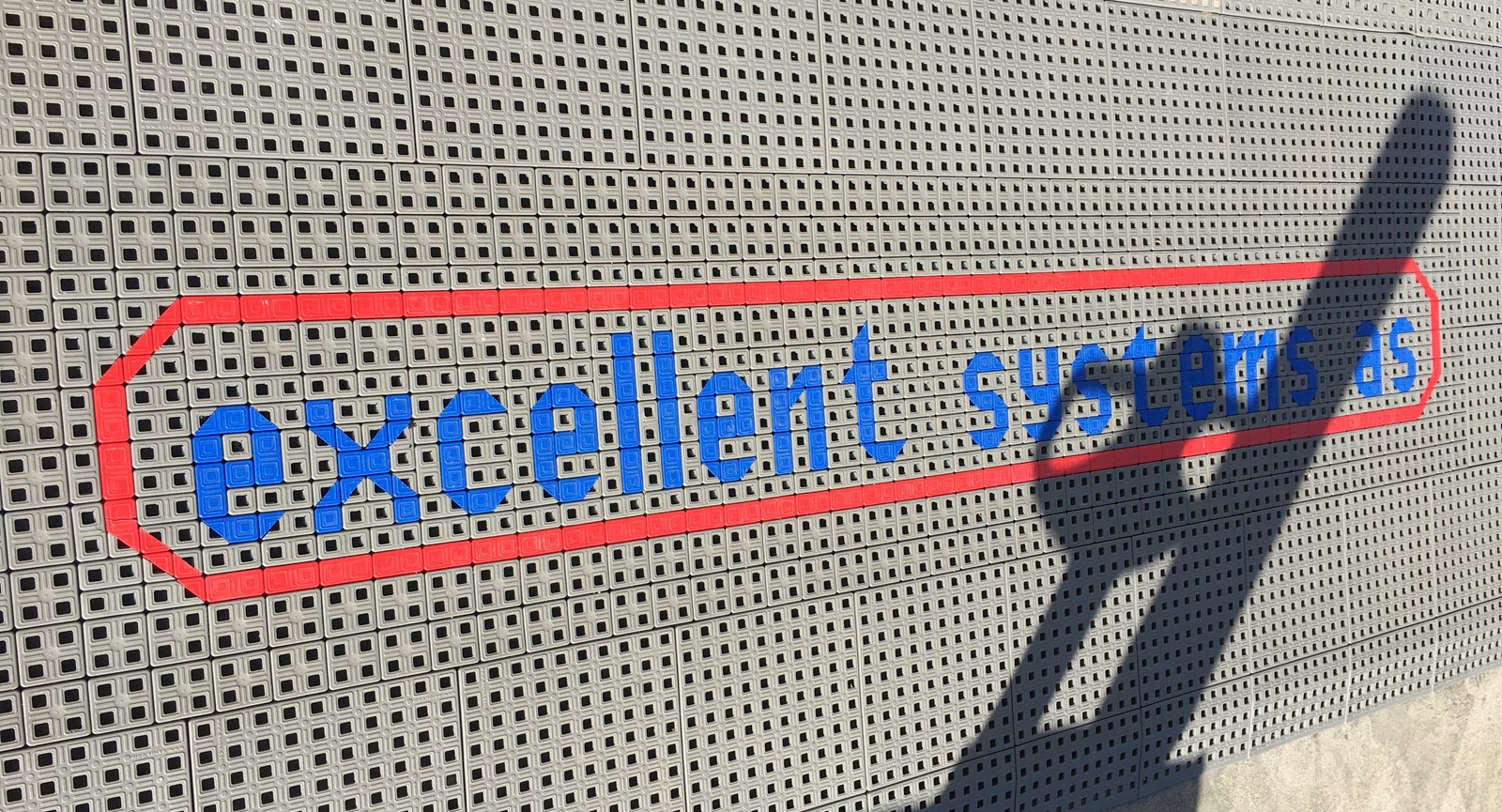 Et aflangt stykke gråt skridsikkert gulv med firmanavnet 'Excellent Systems' skrevet med lyseblå bogstaver.