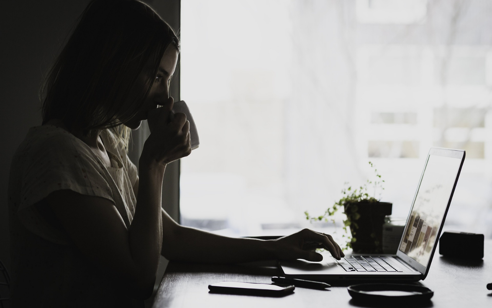 Kvinde sidder ved sin computer og drikker af et krus med en hånd på sin pc