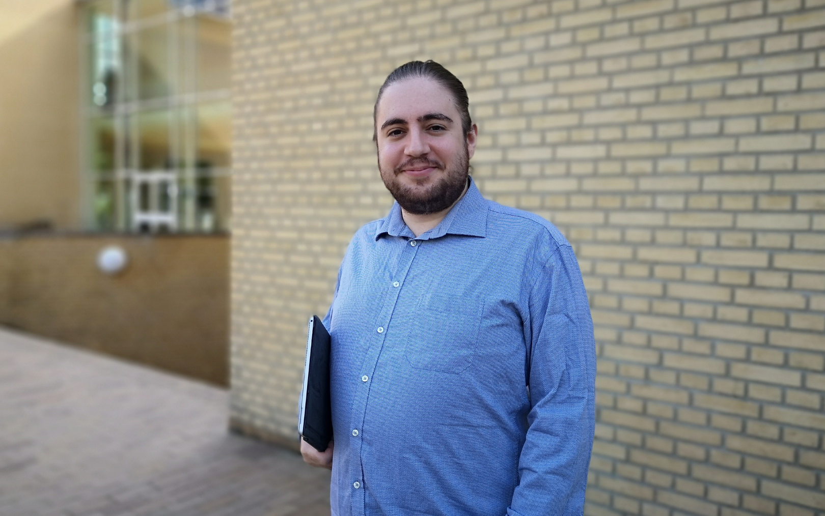 Jacob Mattar Olsson smiler foran Aarhus Universitet med sin computer under armen