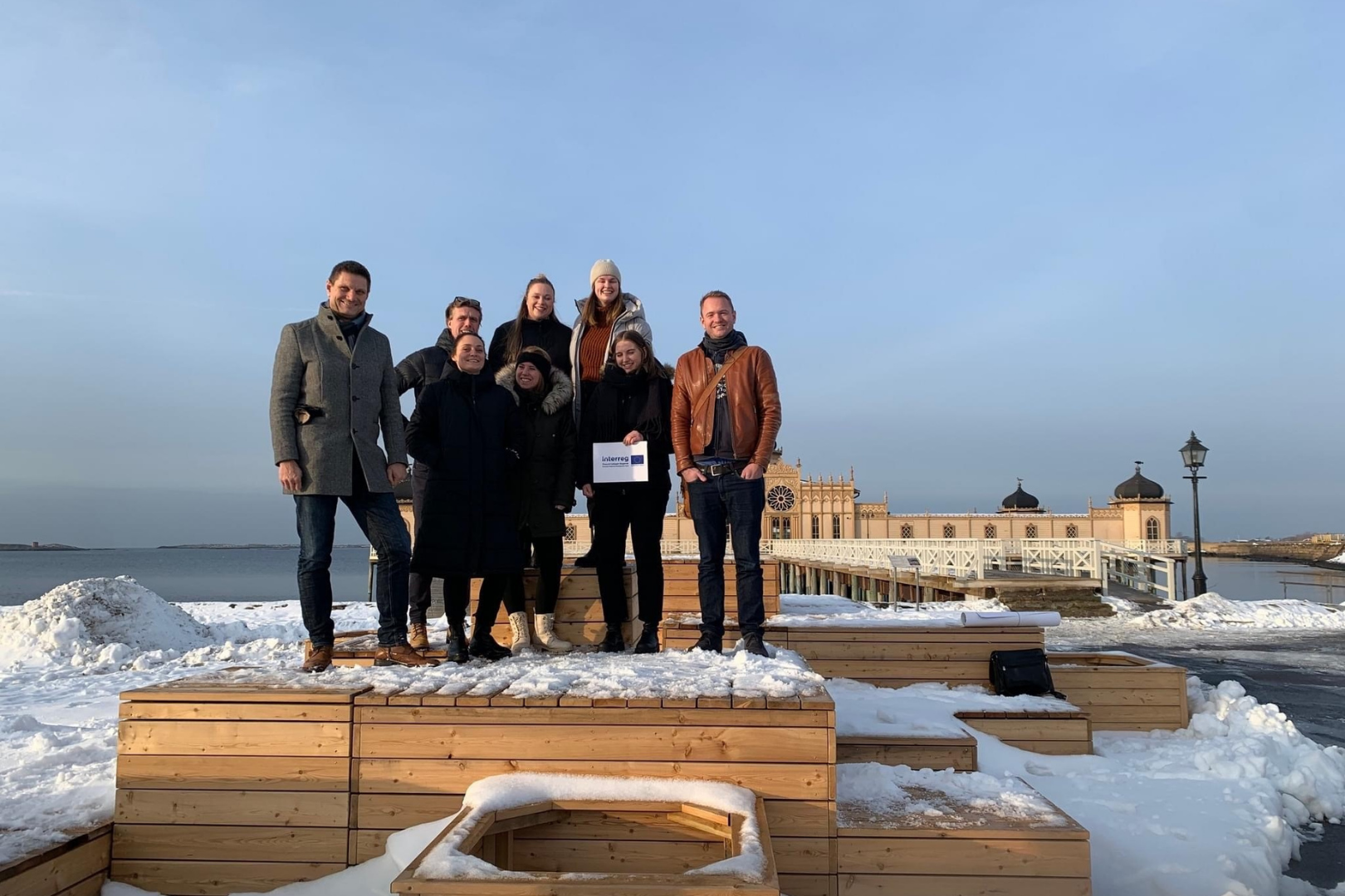 En gruppe af studerende samt 2 projektmedarbejdere står på en trækonstruktion med sne om sig med en svensk badeanstalt i baggrunden i Varberg i Sverige