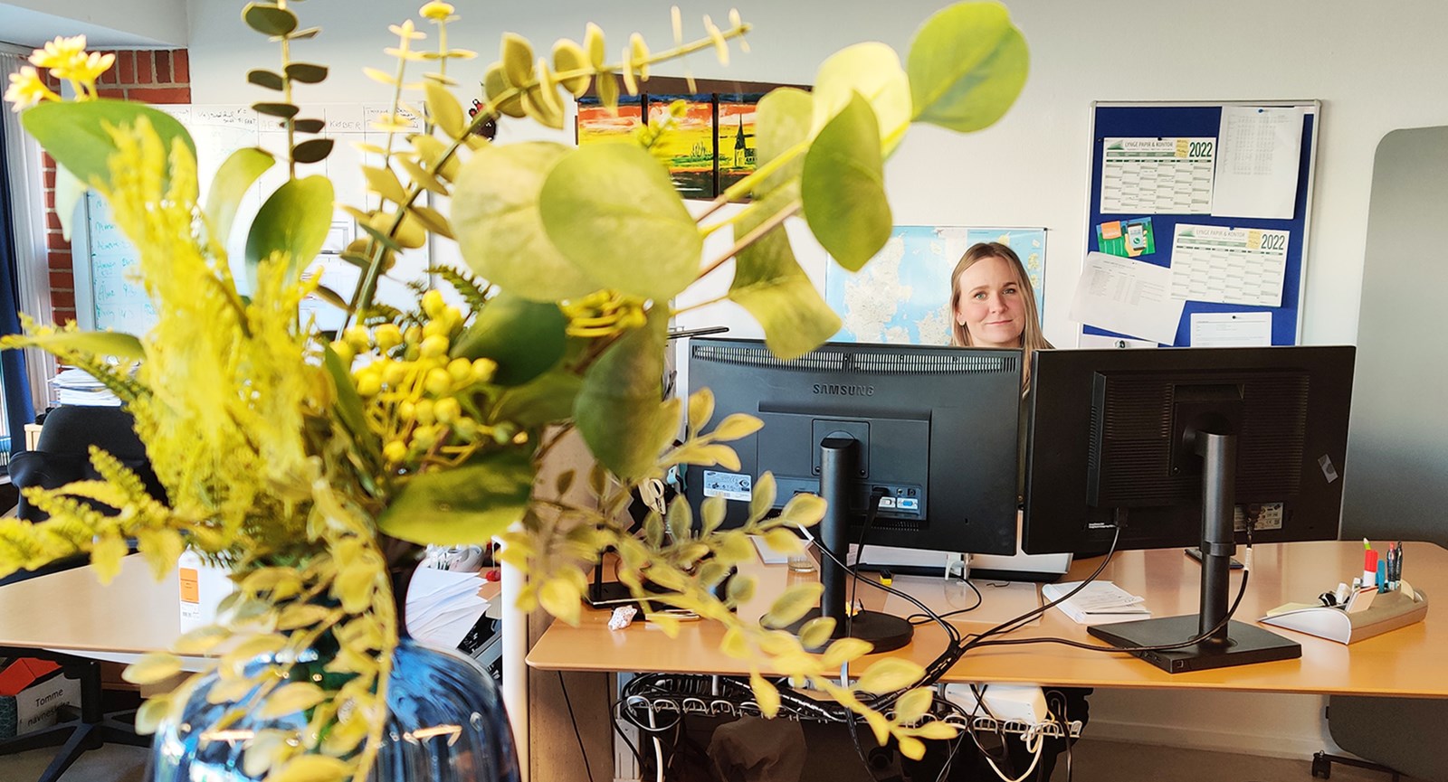 Camilla Østerdal ved computeren i baggrund, mens blomster er i forgrunden