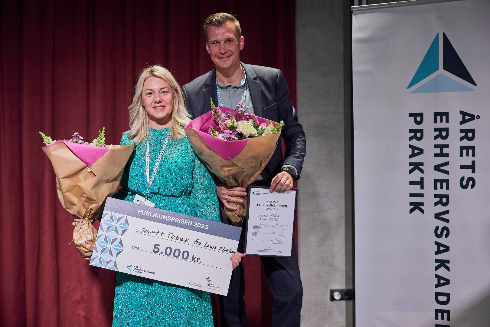 Jeanett, der har læst til optiker på Erhvervsakademi Dania i Randers, står med check og blomster sammen med praktikvejleder Sebastian fra Louis Nielsen efter at have vundet publikumsprisen til Årets Erhvervsakademipraktik 2023