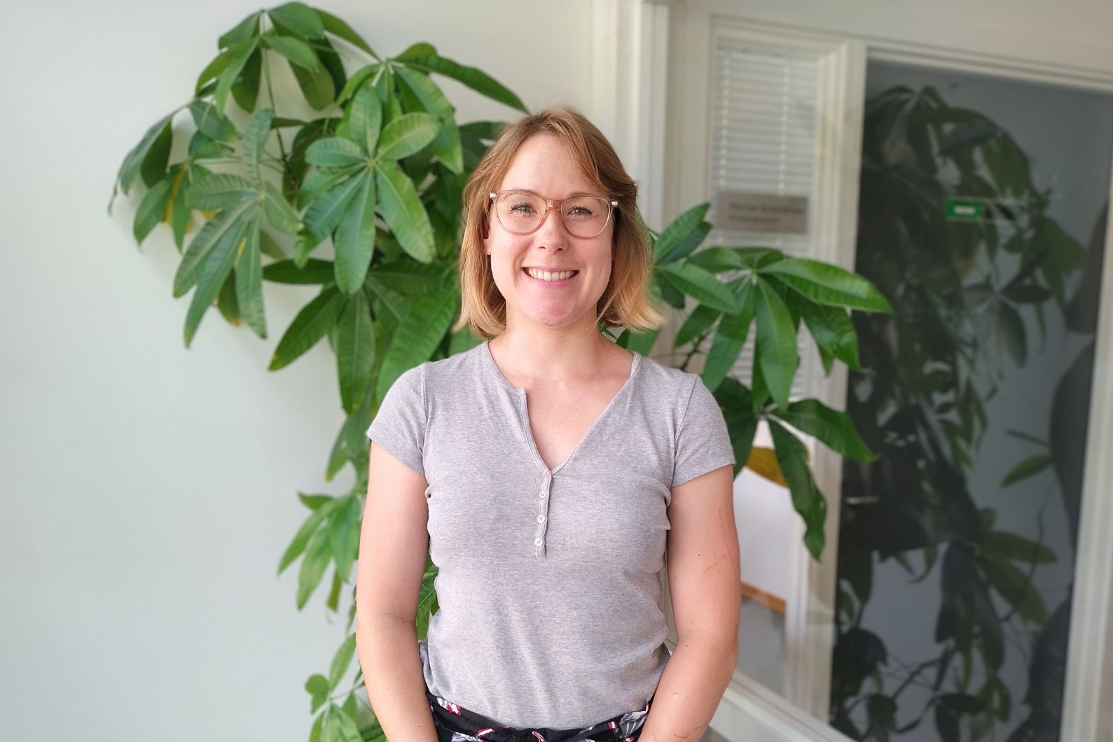 Sara Noer, der er administrationsøkonom fra Erhvervsakademi Dania i Horsens, står foran en grøn plante og smiler til dig