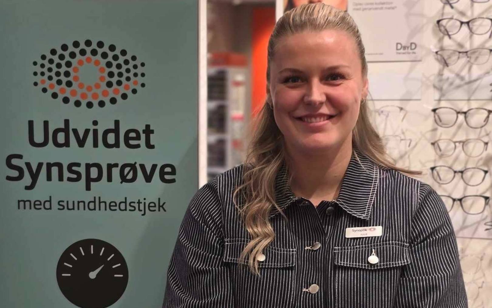Ansøger Julie til optometri studiet online smiler til kameraet med Synoptik skilt i baggrunden