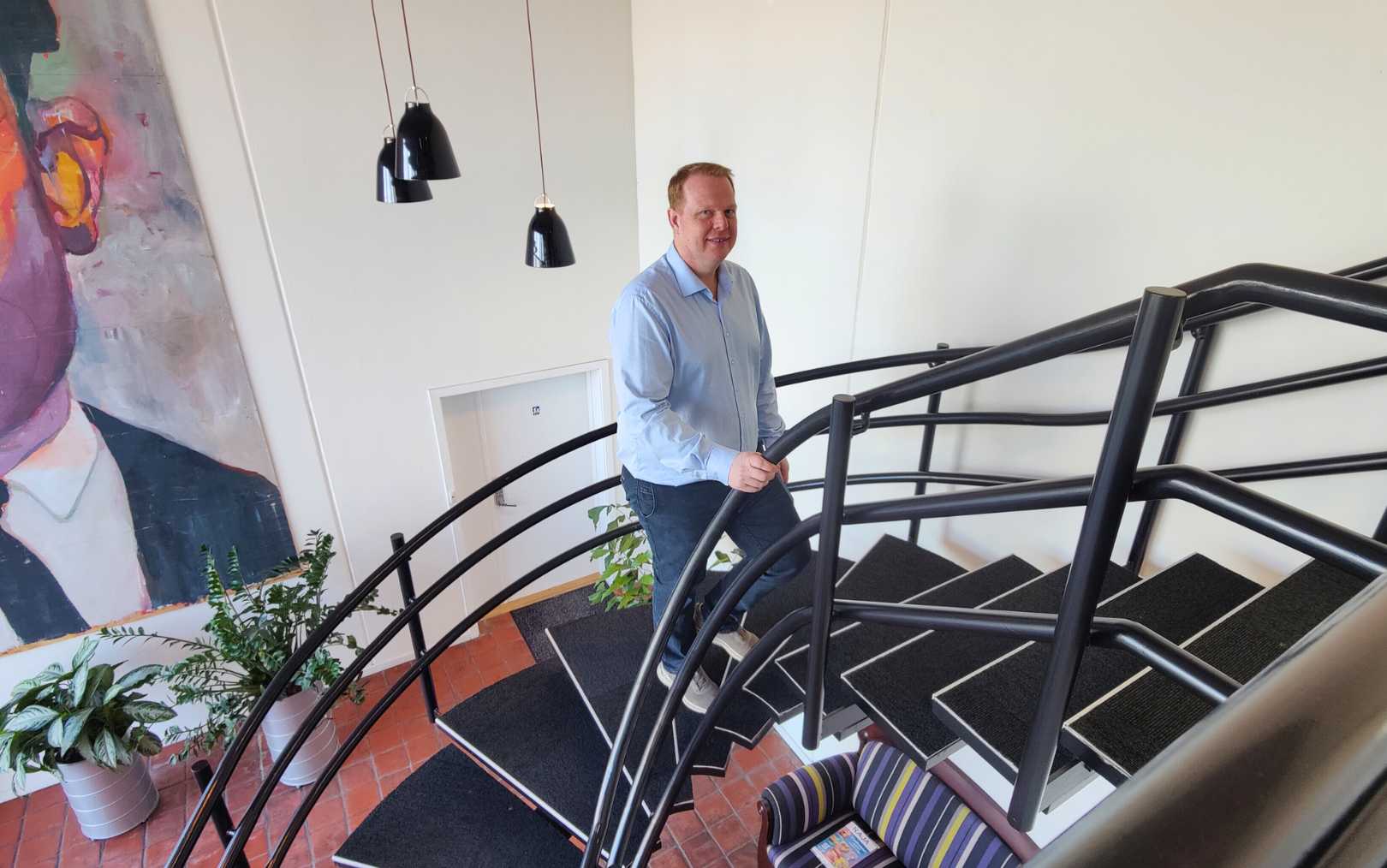 Jens Lindskov Vestergaard er afdelingschef ved Holtec og uddannet automationsteknolog på Erhvervsakademi Dania. Han står på en trappe med et stort maleri i baggrunden og sorte lamper i baggrunden