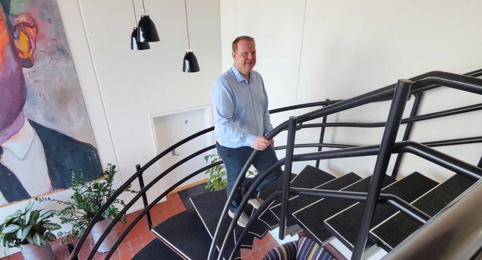 Jens Lindskov Vestergaard er afdelingschef ved Holtec og uddannet automationsteknolog på Erhvervsakademi Dania. Han står på en trappe med et stort maleri i baggrunden og sorte lamper i baggrunden