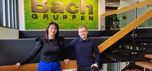 Camilla og Peter står foran et grønt skilt med Bach Gruppens logo, da de er i finalen som Erhvervsakademi Danias bedste praktikophold