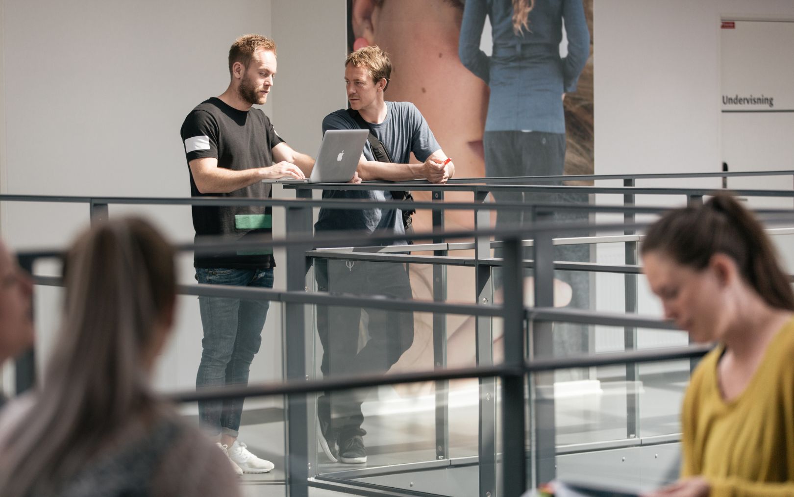 2 studerende fra Erhvervsakademi Dania står på gangen og snakker med deres computer, mens 2 studerende ses i forgrunden